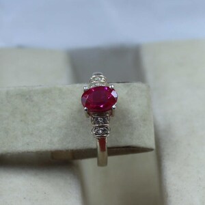 May include: A silver ring with a large, oval-cut ruby gemstone. The ruby is a deep red colour. The ring's band is decorated with smaller, clear gemstones on either side of the ruby, adding sparkle to the design.