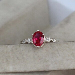 May include: A silver ring featuring a large, oval-cut, vibrant red gemstone as the centerpiece. The band is adorned with smaller, clear gemstones and decorative accents. The ring is set against a neutral background, highlighting its elegant design.
