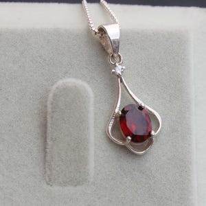 Könnte beinhalten: Eine silberne Anhängerkette mit einem tiefroten, ovalen Edelstein. Der Anhänger hat ein filigranes, offenes Design mit einem kleinen, klaren Akzentstein über dem Hauptstein. Die Halskette ist ebenfalls aus Silber.
