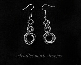 May include: A pair of silver dangle earrings with multiple interconnected rings. The earrings are on a black background. @feuilles.morte.designs