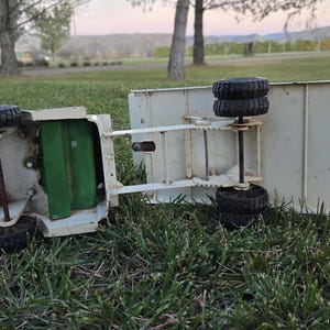 May include: An overturned vintage toy truck with a cream-colored body and black tires. The truck's bed is rectangular, and the wheels are visible. The truck is lying on green grass, with a green interior visible. The background shows trees and a field.