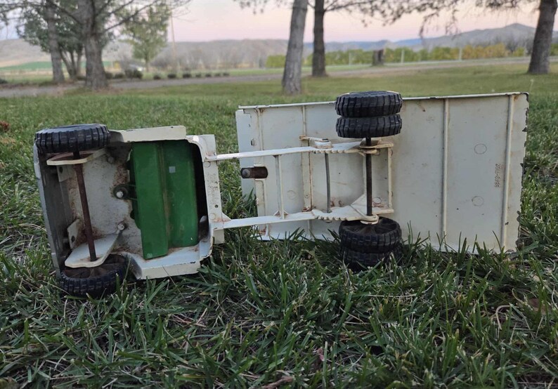 May include: An overturned vintage toy truck with a cream-colored body and black tires. The truck's bed is rectangular, and the wheels are visible. The truck is lying on green grass, with a green interior visible. The background shows trees and a field.