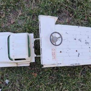 May include: A vintage white toy truck with a flatbed and a cab. The truck has a steering wheel and a sticker that reads "TO OPERATE TURN WHEEL". The toy is sitting on a bed of green grass.