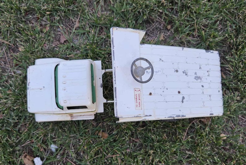 May include: A vintage white toy truck with a flatbed and a cab. The truck has a steering wheel and a sticker that reads "TO OPERATE TURN WHEEL". The toy is sitting on a bed of green grass.