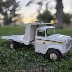 May include: A vintage white and green toy truck with a flatbed, black tires, and a John Deere logo. The toy truck is made of metal and is sitting on green grass. The truck has a yellow stripe along the side.