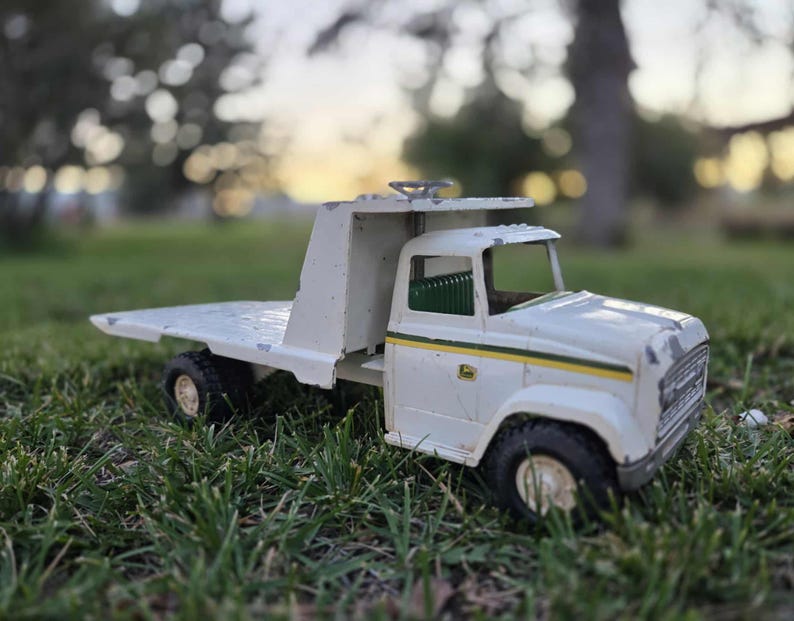 May include: A vintage white and green toy truck with a flatbed, black tires, and a John Deere logo. The toy truck is made of metal and is sitting on green grass. The truck has a yellow stripe along the side.