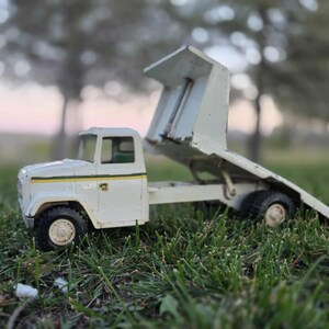 May include: A vintage white toy dump truck with a raised bed, set against a blurred background of trees and a dusky sky. The truck has black tires and a green and yellow stripe detail. The toy is resting on a bed of green grass.