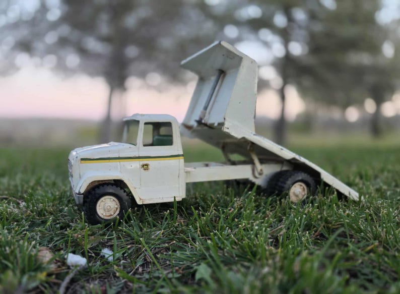 May include: A vintage white toy dump truck with a raised bed, set against a blurred background of trees and a dusky sky. The truck has black tires and a green and yellow stripe detail. The toy is resting on a bed of green grass.