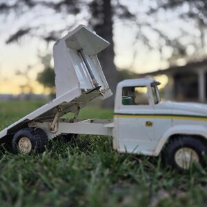 May include: A vintage white toy dump truck with a raised bed, set on green grass. The truck has black tires, a yellow stripe, and a small cab. The bed is angled upwards, showcasing its interior.