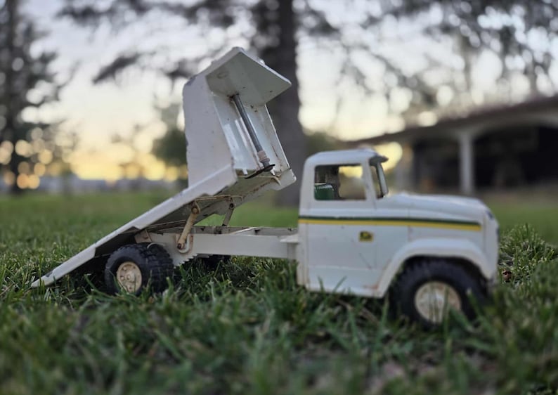 May include: A vintage white toy dump truck with a raised bed, set on green grass. The truck has black tires, a yellow stripe, and a small cab. The bed is angled upwards, showcasing its interior.