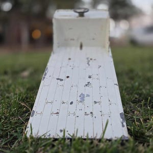 May include: A weathered, white metal ramp with a handle on top, set on a grassy surface. The ramp has a rectangular shape with a series of parallel lines and visible wear and tear, suggesting age and use. The background is blurred.