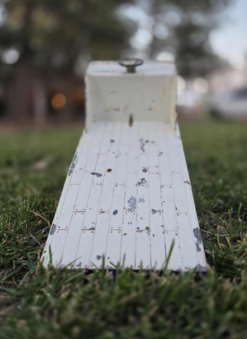 May include: A weathered, white metal ramp with a handle on top, set on a grassy surface. The ramp has a rectangular shape with a series of parallel lines and visible wear and tear, suggesting age and use. The background is blurred.