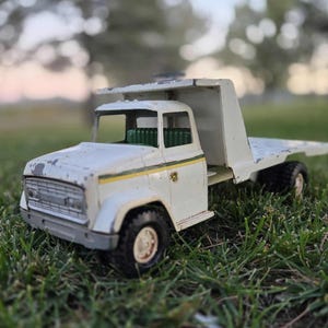 May include: A vintage white toy truck with a flatbed, set against a blurred green background. The truck has black tires, a green interior, and yellow and green stripes. The paint shows signs of wear.