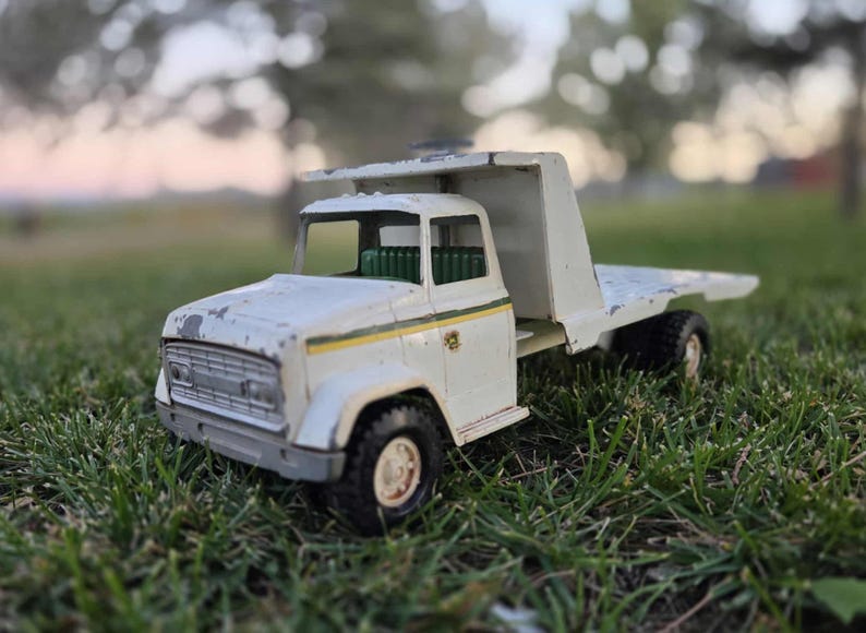 May include: A vintage white toy truck with a flatbed, set against a blurred green background. The truck has black tires, a green interior, and yellow and green stripes. The paint shows signs of wear.