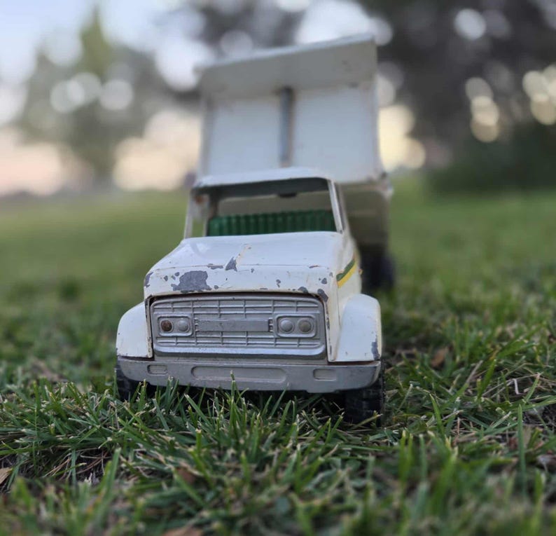 May include: A vintage white toy dump truck with a raised bed, showing signs of wear. The truck has a silver grill, black tires, and a green interior. The background is blurred, with green grass in the foreground.