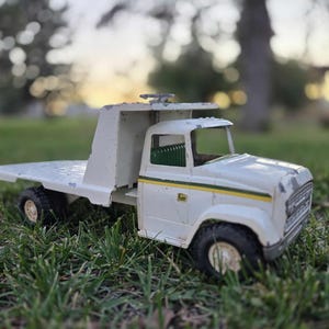 May include: A vintage white toy truck with a flatbed, black tires, and a green and yellow stripe detail. The toy truck is sitting on green grass.