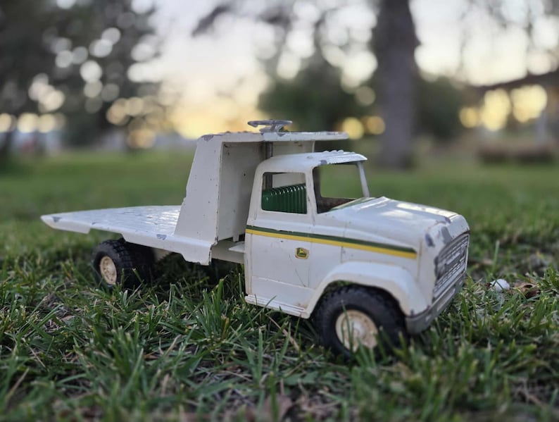 May include: A vintage white toy truck with a flatbed, black tires, and a green and yellow stripe detail. The toy truck is sitting on green grass.