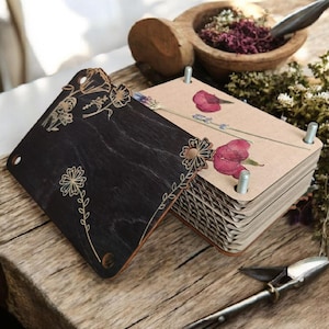 May include: A wooden flower press with a dark stained top and etched floral designs. The press is open, revealing pages with pressed flowers. A wooden bowl with herbs and a metal tool are in the background, suggesting a botanical theme.