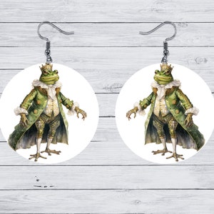 Puede incluir: Un par de pendientes redondos blancos con una rana verde que lleva una corona dorada y un abrigo estampado en verde y blanco. La rana está de pie sobre un fondo blanco.