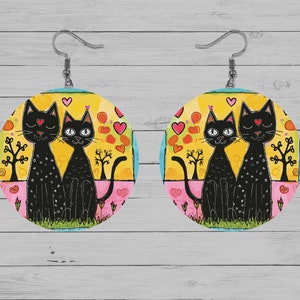 May include: A pair of round, colourful earrings featuring a black cat couple with hearts and flowers. The earrings are made of wood and have a silver hook.