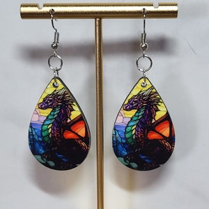 May include: A pair of teardrop-shaped earrings with a stained glass dragon design. The dragon is purple, green, and orange with a yellow background. The earrings are made of wood and have silver hooks.