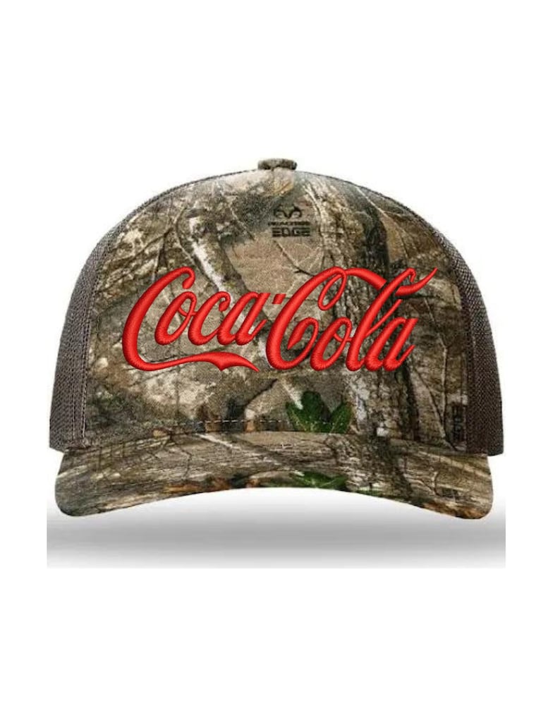 May include: A brown and green camouflage trucker hat with a red embroidered Coca-Cola logo. The hat has a black mesh back and a white "Wilderness Edge" logo.