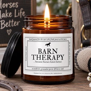 Op de afbeelding: Een brandende kaars in een amberkleurige glazen pot met een zwarte deksel. Het witte etiket toont een zwart silhouet van een paard, de tekst BARN THERAPY, de zin because horses listen better en informatie over handgegoten sojawas.