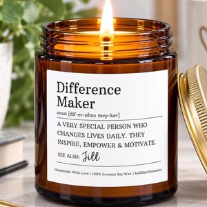 May include: A lit candle in an amber glass jar with a white label featuring the text Difference Maker, a dictionary-style definition, and the name Jill. The label includes the text Handmade With Love, 100% Coconut Soy Wax, and KaliBayElements.
