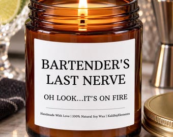 May include: A lit candle in an amber glass jar with a white label reading BARTENDER'S LAST NERVE, OH LOOK...IT'S ON FIRE, Handmade With Love | 100% Natural Soy Wax | KaliBayElements. The candle features a visible flame and a gold-coloured lid.