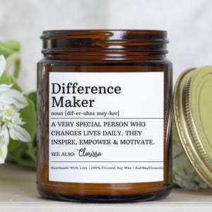 May include: A brown glass jar candle with a gold lid. The label reads "Difference Maker" with a definition and inspirational text. The candle is surrounded by white flowers and green foliage.