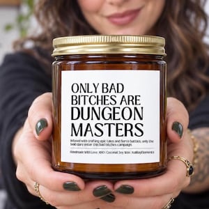 May include: A brown glass candle jar with a gold metal lid and a white rectangular label. The label features the black text: ONLY BAD BITCHES ARE DUNGEON MASTERS, followed by smaller text about epic tales, fierce battles, and ingredients.