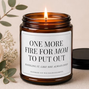 May include: A lit brown glass candle with a black lid. The candle has a white label with the text "ONE MORE FIRE FOR MOM TO PUT OUT. HANDLING IT, LIKE SHE ALWAYS DOES!" The candle is handmade with soy wax.