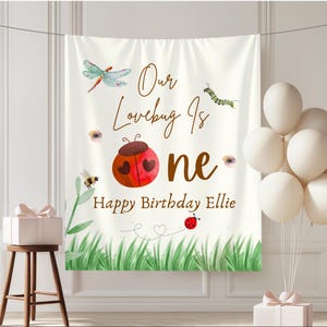 May include: A white fabric banner with the text "Our Lovebug Is One Happy Birthday Ellie". The banner features illustrations of a ladybug, dragonfly, bee, and caterpillar, with green grass at the bottom and white balloons.