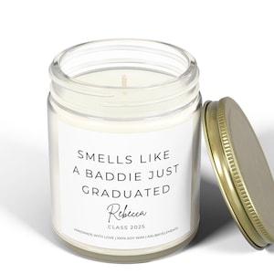 A white candle jar with a gold lid. The label reads "Smells like a baddie just graduated" with the name "Rebecca" and "Class 2025" underneath. The candle is handmade with 100% soy wax.