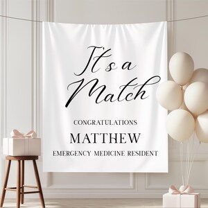 Puede incluir: Bandera blanca con texto negro "It's a Match". Debajo, "Congratulations Matthew Emergency Medicine Resident". La imagen incluye globos blancos y una caja de regalo, creando un ambiente festivo.