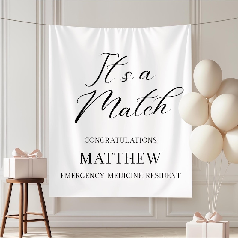 Puede incluir: Bandera blanca con texto negro "It's a Match". Debajo, "Congratulations Matthew Emergency Medicine Resident". La imagen incluye globos blancos y una caja de regalo, creando un ambiente festivo.