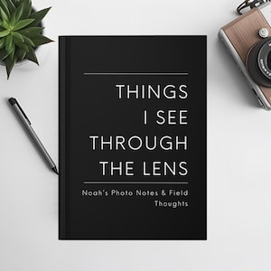 May include: A black notebook with the text "THINGS I SEE THROUGH THE LENS" and "Noah's Photo Notes & Field Thoughts" in white. A silver pen, a succulent plant, and a vintage camera with a brown leather strap are also visible.