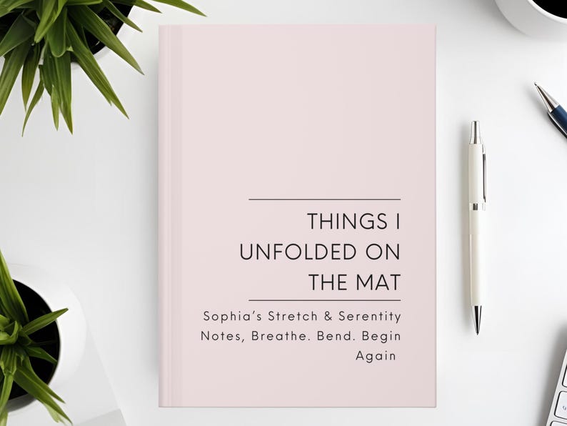 以下が含まれることがあります： 「THINGS I UNFOLDED ON THE MAT」というタイトルの薄いピンク色のジャーナル。テキストは「Sophia's Stretch & Serenity Notes, Breathe. Bend. Begin Again」と書かれています。白いペンとコーヒーカップが白い表面にあります。