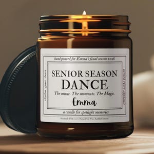 May include: A brown glass candle jar with a black lid, featuring a white label with the text "SENIOR SEASON DANCE" and "Emma". The candle is lit, and the label includes the phrase "a candle for spotlight memories."