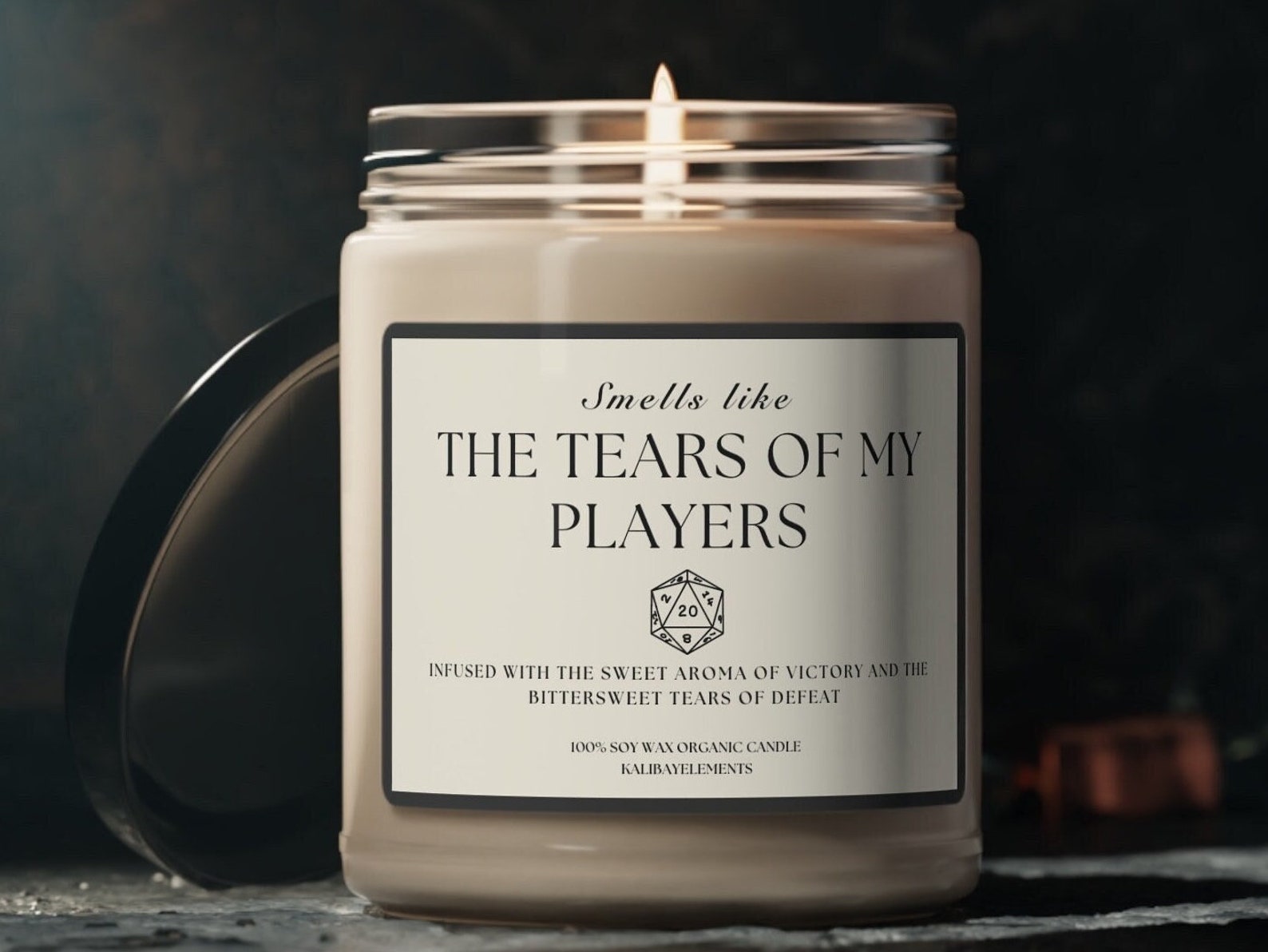 A white jar candle titled "Smells like the tears of my players" is lit on a table with the lid leaning up against it.