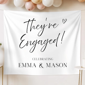 Può includere: Banner di fidanzamento bianco con le parole "They're Engaged!" in elegante scrittura nera, un piccolo cuore e i nomi Emma & Mason. Lo striscione è decorato con palloncini e una torta.