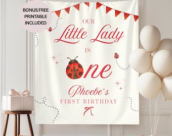 May include: A cream-colored backdrop with red text and ladybug illustrations. The text reads "Our Little Lady is One Phoebe's First Birthday." A string of red pennants and white balloons are also visible.