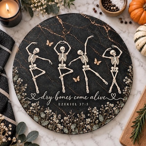 May include: A round black serving board featuring four dancing skeletons, small butterflies, and white floral accents. The text reads "dry bones come alive" and "EZEKIEL 37:5" in white script and block lettering against a dark marble-like background.