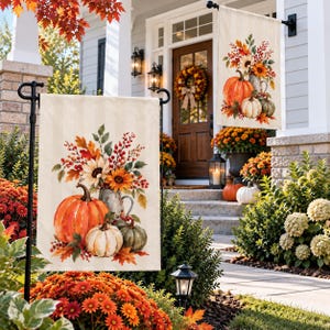May include: A rectangular garden flag featuring a printed illustration of three pumpkins in orange, white, and green, arranged beside a metal pitcher filled with sunflowers, autumn leaves, and red berries, all set against a light cream background.