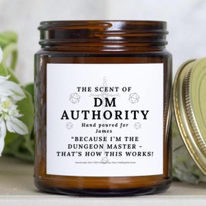 May include: A brown glass jar candle with a white label. The label reads "THE SCENT OF DM AUTHORITY" and "Hand poured for James." Below, it says "BECAUSE I'M THE DUNGEON MASTER - THAT'S HOW THIS WORKS!" Handmade in the USA.