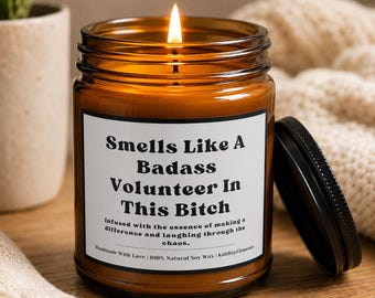 Pode incluir: Uma vela acesa num frasco de vidro âmbar com um rótulo branco com o texto preto: Smells Like A Badass Volunteer In This Bitch. Infused with the essence of making a difference and laughing through the chaos. Handmade With Love | 100% Natural Soy Wax | kaliBayElements. Uma tampa de metal preta está ao lado do frasco.