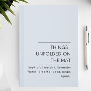 以下が含まれることがあります： 「THINGS I UNFOLDED ON THE MAT」と書かれた水色のノート。タイトル下には「Sophia's Stretch & Serenity Notes, Breathe. Bend. Begin Again」の文字。白いペンとコーヒーカップが白い表面に置かれています。