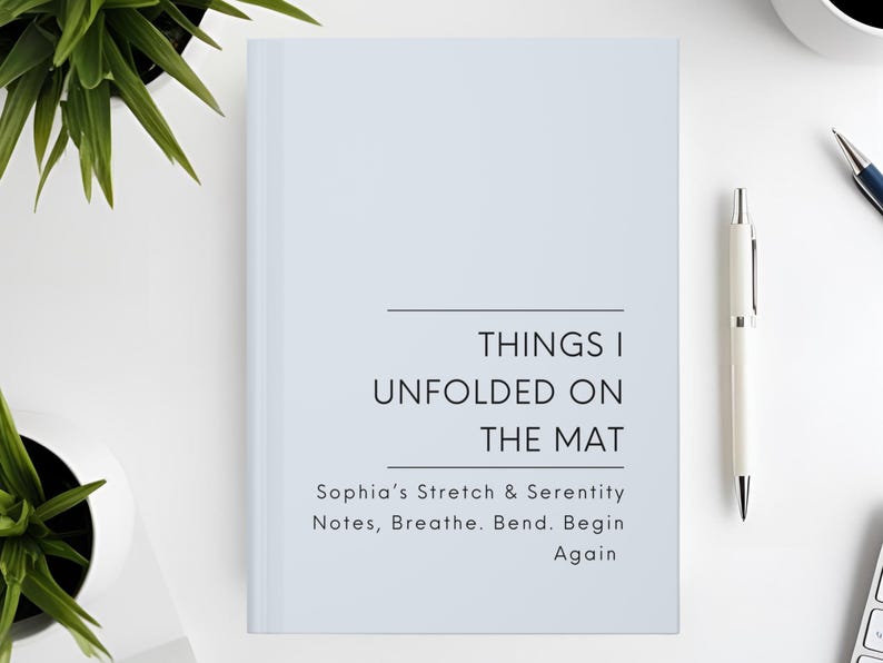 以下が含まれることがあります： 「THINGS I UNFOLDED ON THE MAT」と書かれた水色のノート。タイトル下には「Sophia's Stretch & Serenity Notes, Breathe. Bend. Begin Again」の文字。白いペンとコーヒーカップが白い表面に置かれています。