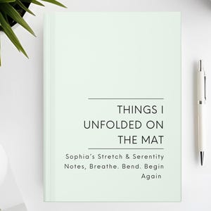 以下が含まれることがあります： 「THINGS I UNFOLDED ON THE MAT」と「Sophia's Stretch & Serenity Notes, Breathe. Bend. Begin Again」の文字が入った、薄い緑色のノート。白いペンとコーヒーカップが白い表面に置かれています。