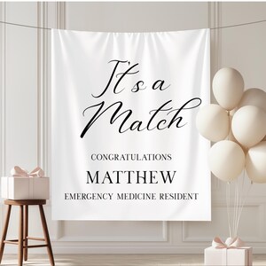 Puede incluir: Bandera blanca con texto negro "It's a Match". Debajo, "Congratulations Matthew Emergency Medicine Resident". La imagen incluye globos blancos, una caja de regalo y un peque&ntilde;o taburete de madera.
